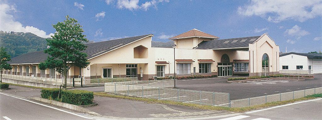 介護施設の全景。クリーム色の平屋建て施設で、駐車場と周囲の緑が見える。